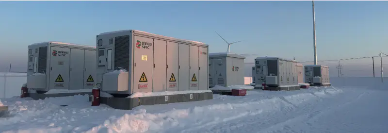 Outdoor power skids and battery enclosures on concrete pads at a snow-covered site, with a wind turbine and transmission line behind them.