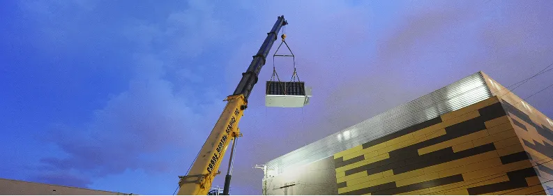 Crane lifting a containerized module onto its pad beside the building.
